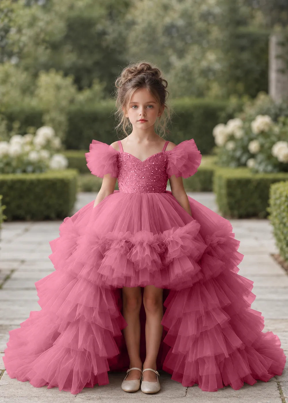 Front view of a child wearing a desert rose flower girl dress with a V-neckline, a sequin-covered bodice and a tiered high-low tulle skirt with off-shoulder sleeve ruffles.