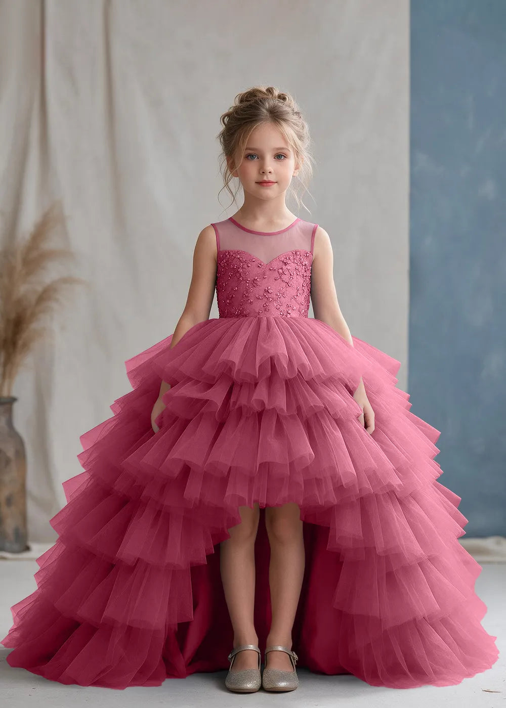 Front view of a child wearing a desert rose flower girl dress with a sheer illusion neckline over a beaded bodice and a high-low tulle skirt with broad tiered ruffle layers.