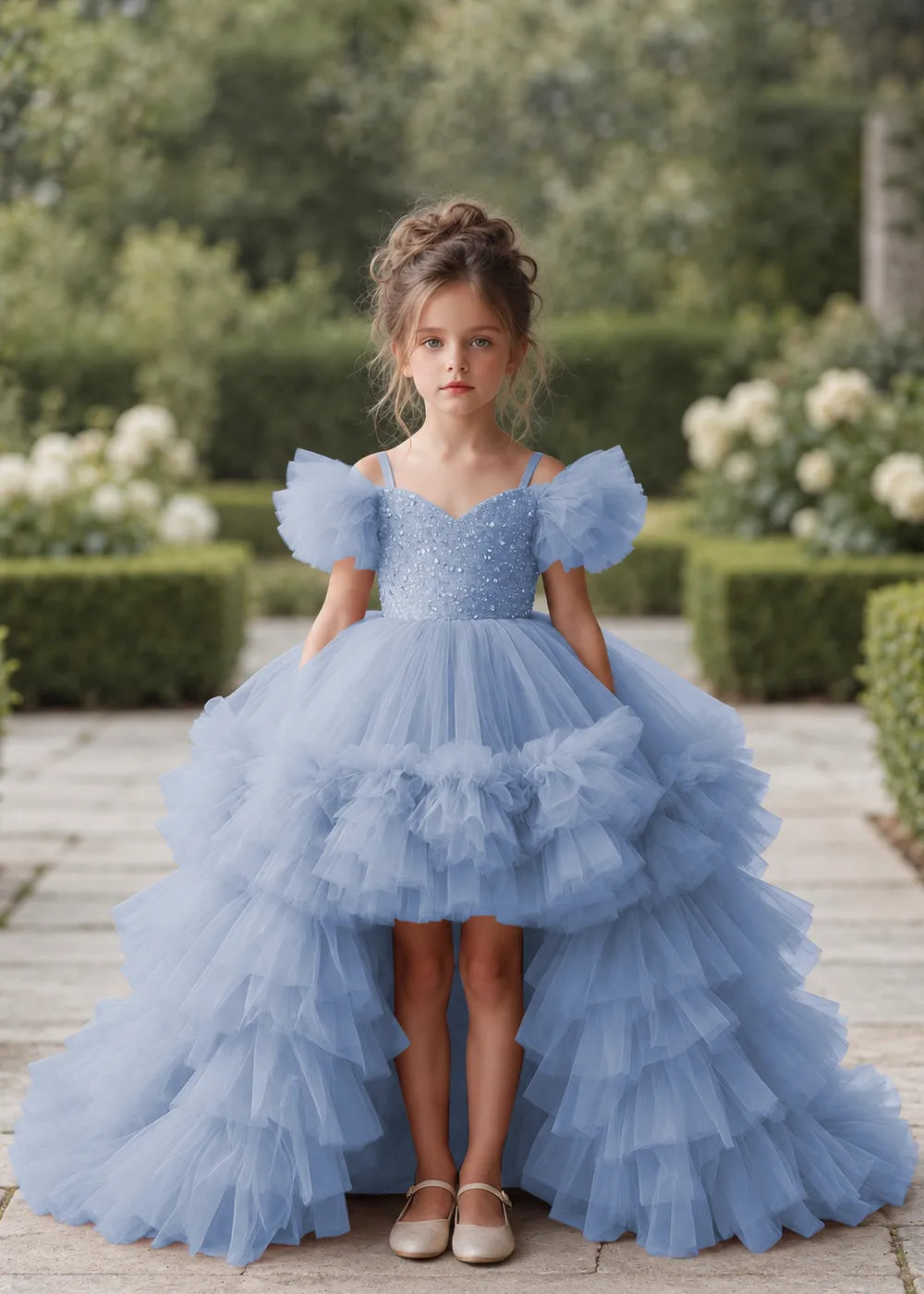 Front view of a child wearing a dusty blue flower girl dress with a V-neckline, a sequin-covered bodice and a tiered high-low tulle skirt with off-shoulder sleeve ruffles.