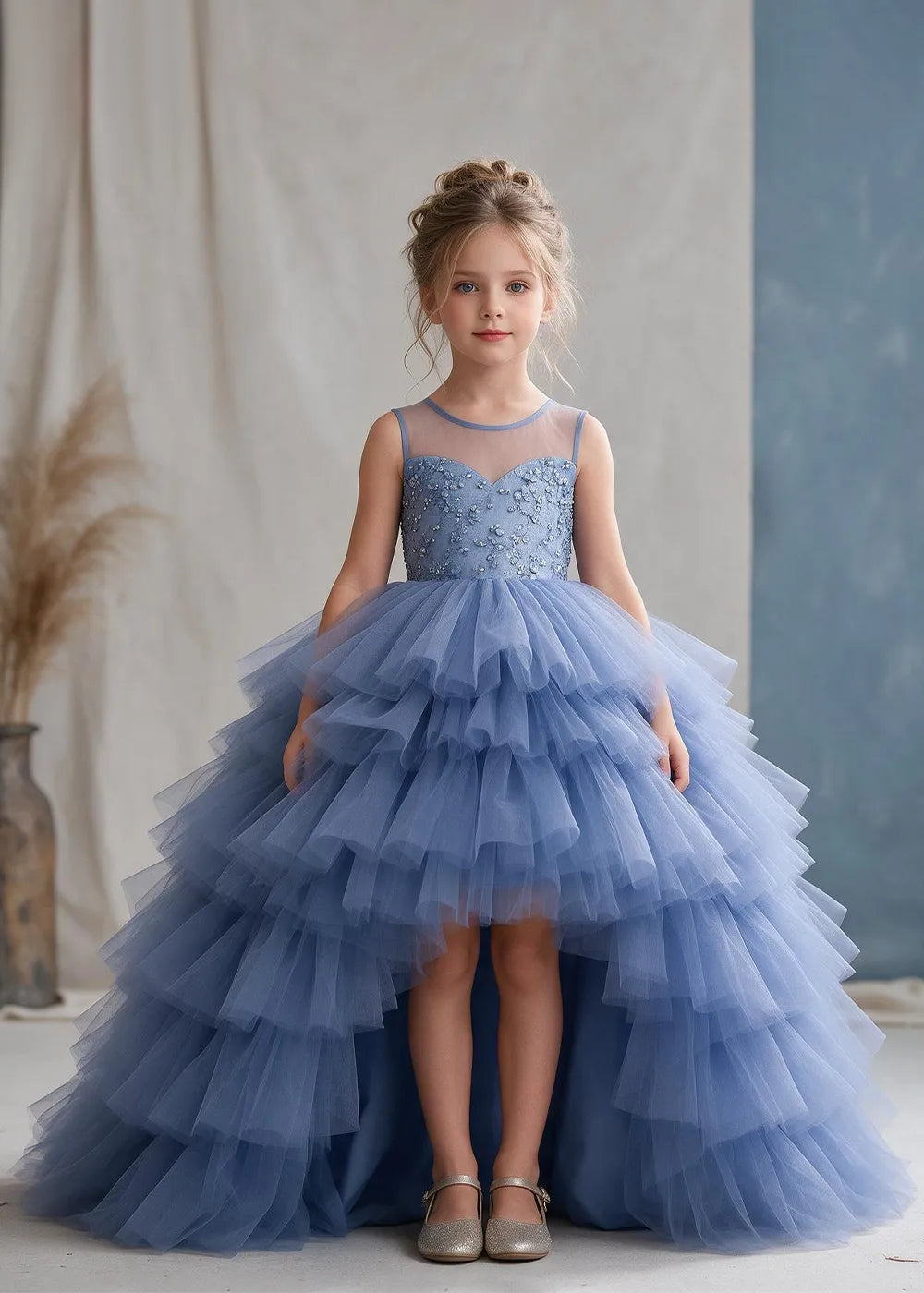 The front view of a child wearing a Dusty Blue flower girl dress with a sheer illusion neckline over a beaded bodice and a high-low tulle skirt with broad tiered ruffle layers.