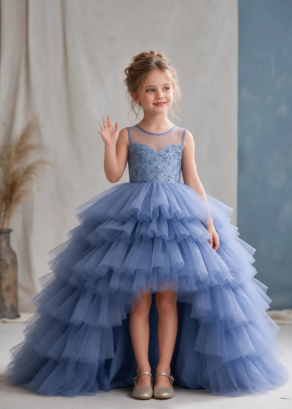 The front view of a child wearing a Dusty Blue flower girl dress with a sheer illusion neckline over a beaded bodice and a high-low tulle skirt with broad tiered ruffle layers.