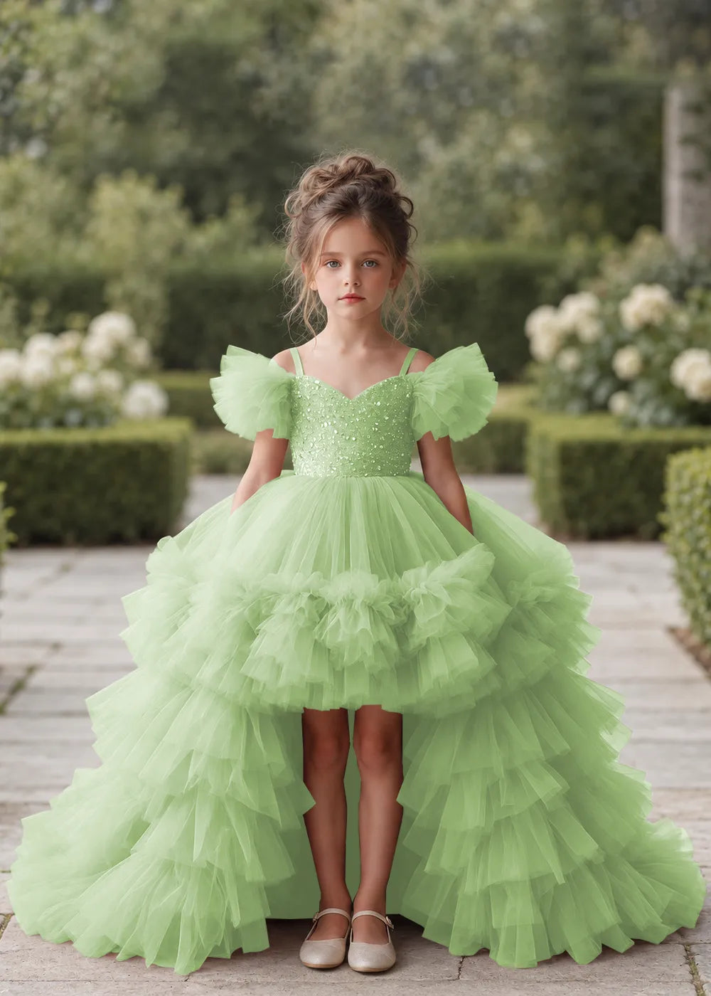 Front view of a child wearing a dusty sage flower girl dress with a V-neckline, a sequin-covered bodice and a tiered high-low tulle skirt with off-shoulder sleeve ruffles.