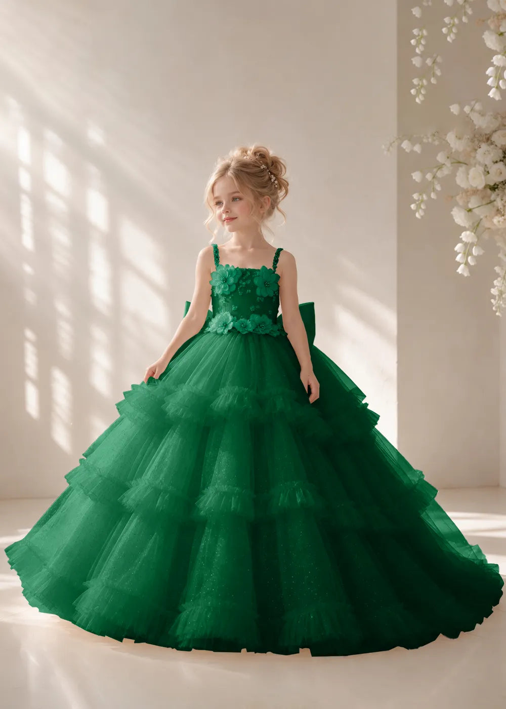 Front view of a child wearing a emerald green flower girl dress with beaded spaghetti straps, a bodice covered in three-dimensional fabric flowers and a multi-tiered tulle skirt.