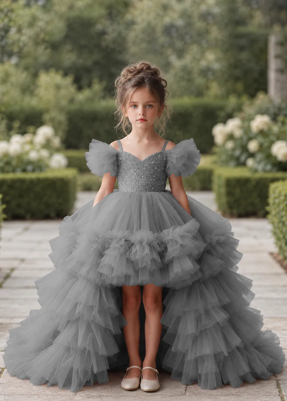 Front view of a child wearing a grey flower girl dress with a V-neckline, a sequin-covered bodice and a tiered high-low tulle skirt with off-shoulder sleeve ruffles.
