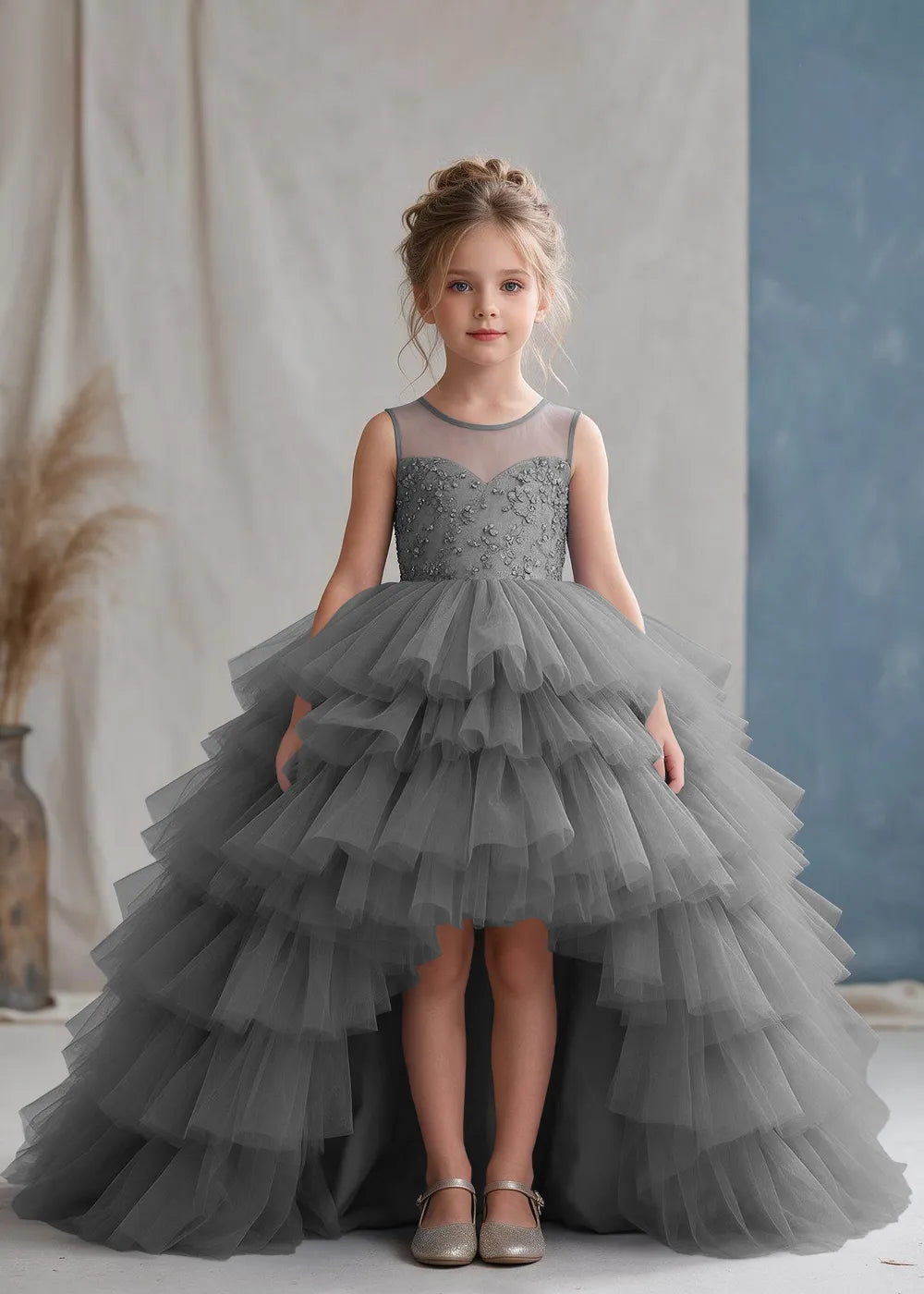 Front view of a child wearing a grey flower girl dress with a sheer illusion neckline over a beaded bodice and a high-low tulle skirt with broad tiered ruffle layers.
