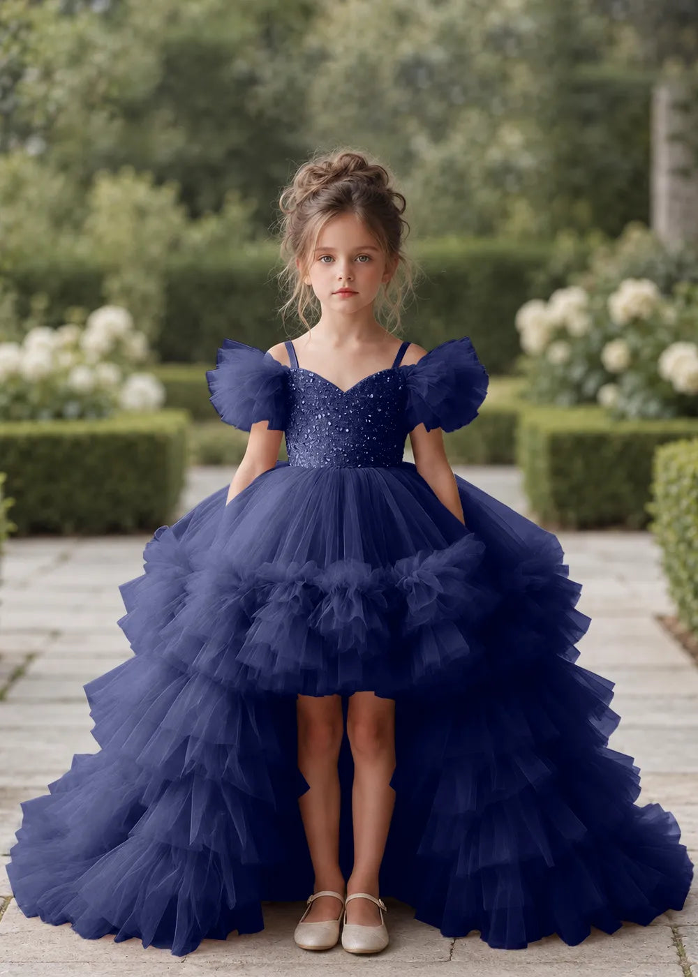 Front view of a child wearing a navy blue flower girl dress with a V-neckline, a sequin-covered bodice and a tiered high-low tulle skirt with off-shoulder sleeve ruffles.