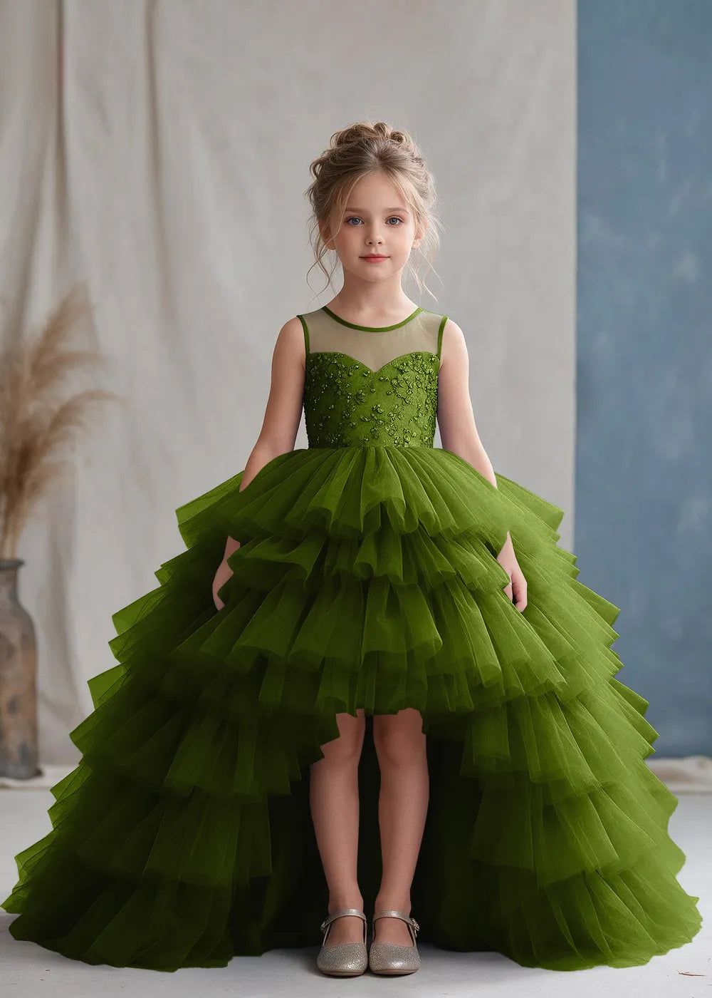 Front view of a child wearing a olive flower girl dress with a sheer illusion neckline over a beaded bodice and a high-low tulle skirt with broad tiered ruffle layers.