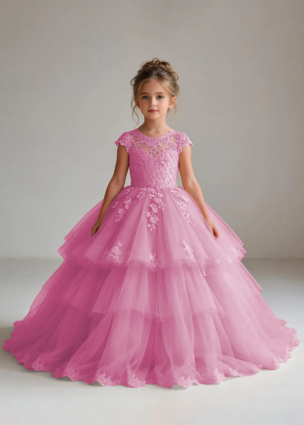 Front view of a child wearing a pearl pink flower girl dress with a sheer lace illusion neckline, cap sleeves and a tiered cake-style tulle skirt with a lace-trimmed hem.