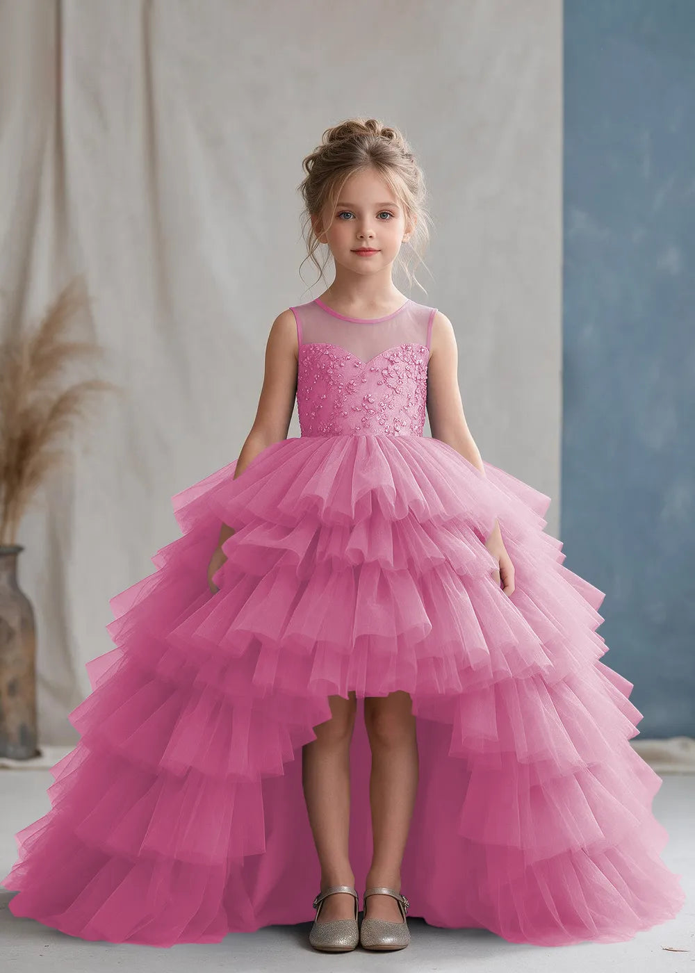Front view of a child wearing a pearl pink flower girl dress with a sheer illusion neckline over a beaded bodice and a high-low tulle skirt with broad tiered ruffle layers.