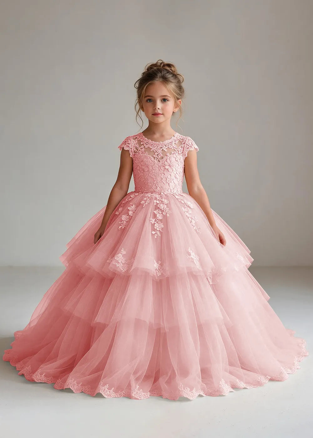 Front view of a child wearing a pink flower girl dress with a sheer lace illusion neckline, cap sleeves and a tiered cake-style tulle skirt with a lace-trimmed hem.
