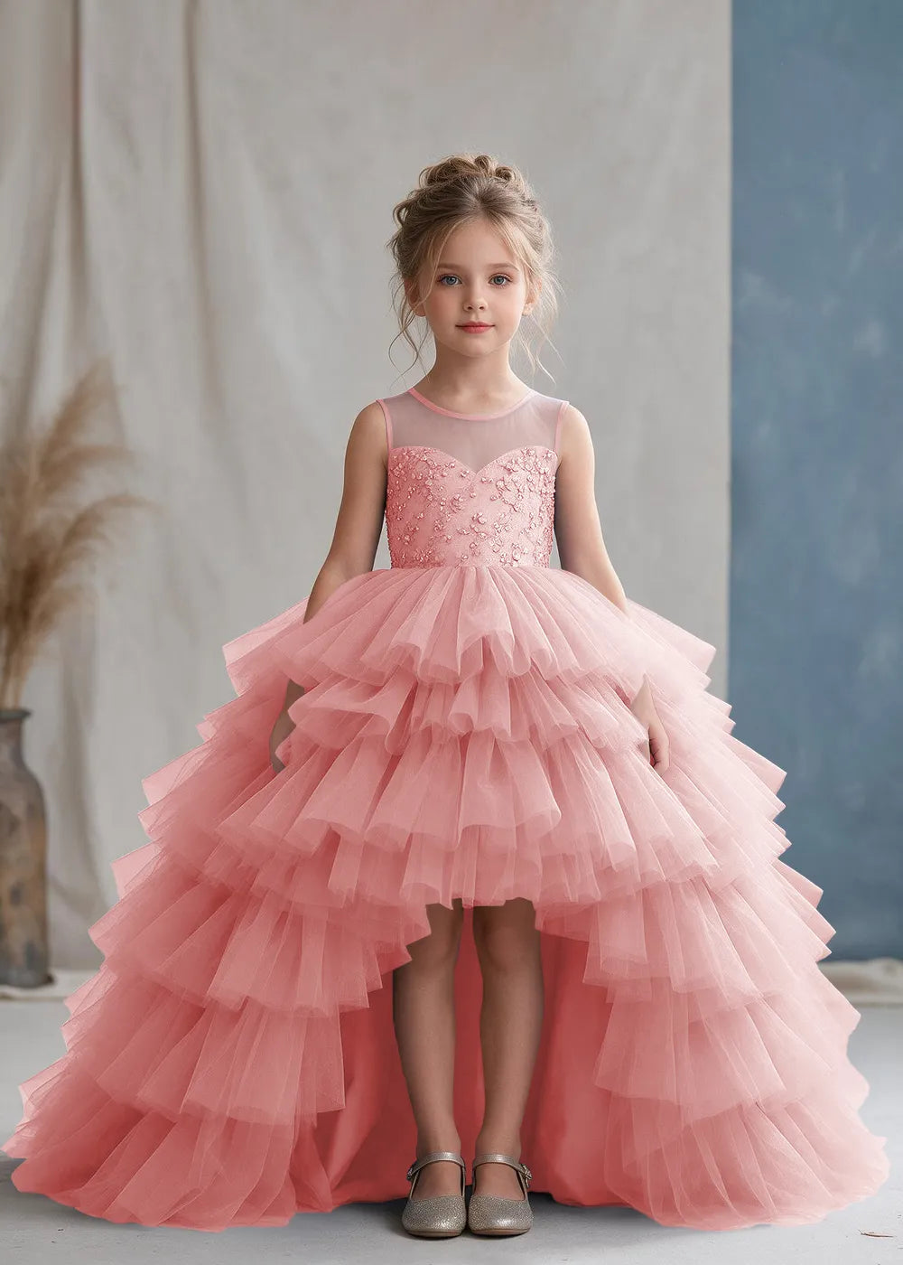 Front view of a child wearing a pink flower girl dress with a sheer illusion neckline over a beaded bodice and a high-low tulle skirt with broad tiered ruffle layers.