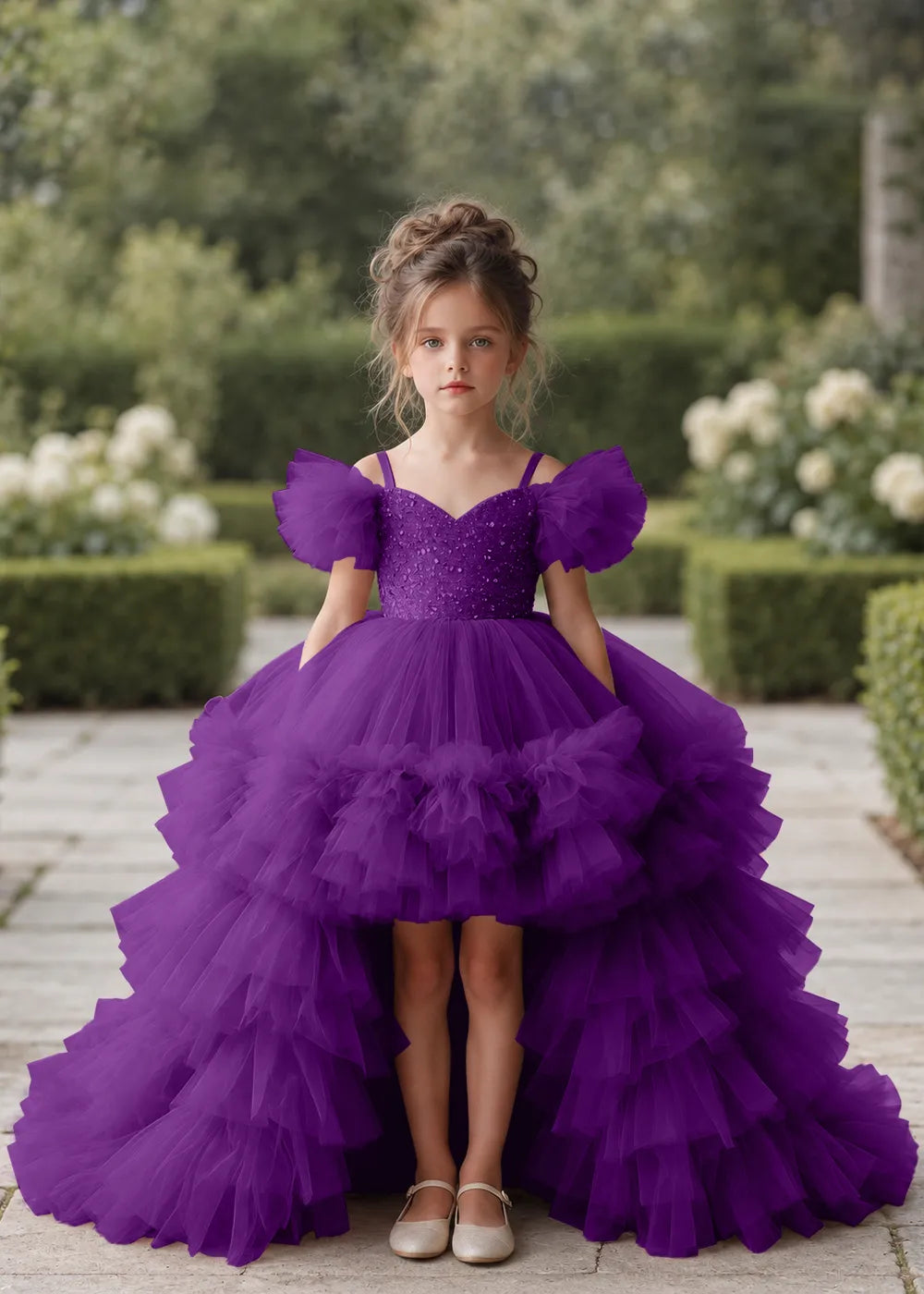 Front view of a child wearing a purple flower girl dress with a V-neckline, a sequin-covered bodice and a tiered high-low tulle skirt with off-shoulder sleeve ruffles.