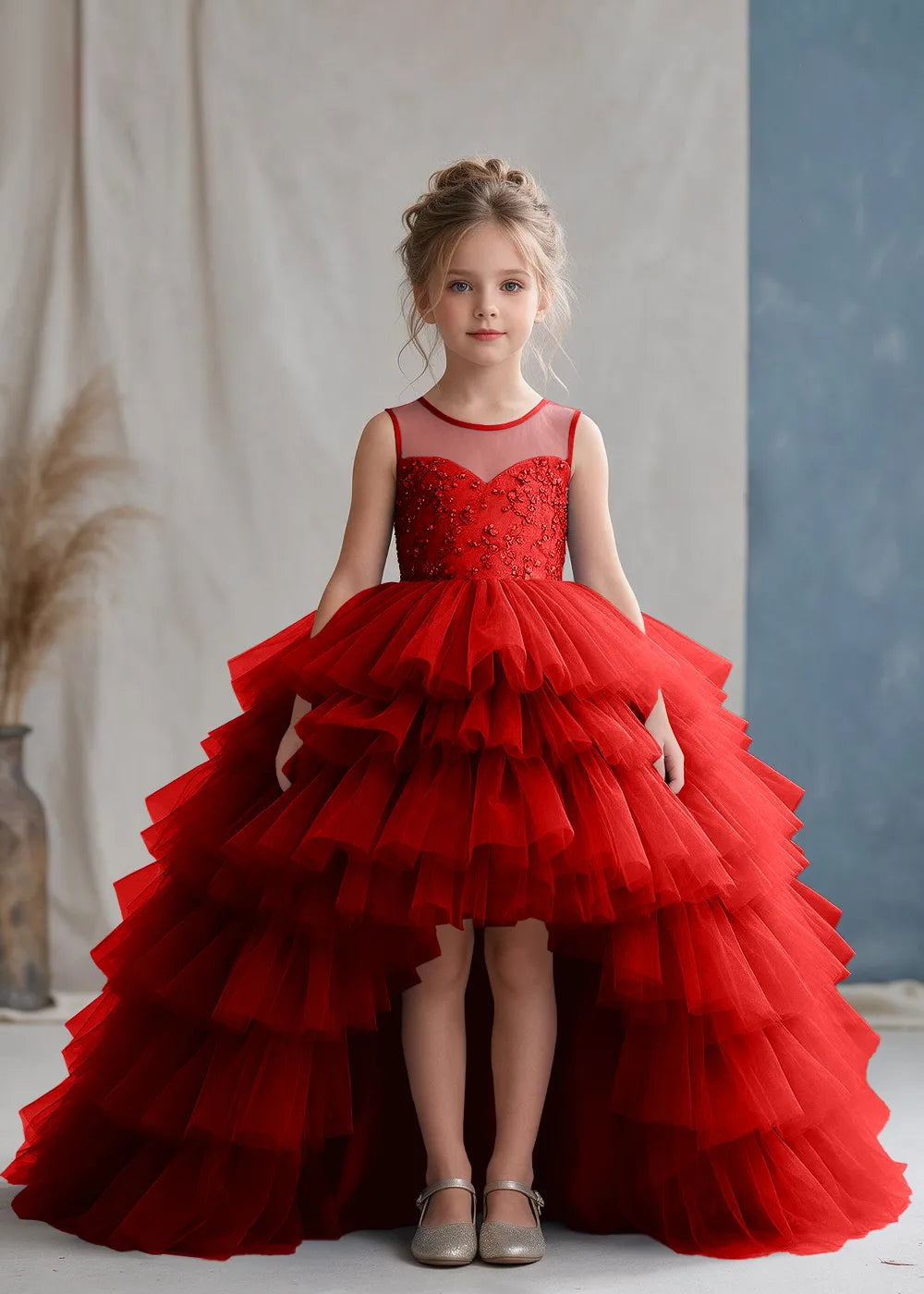 Front view of a child wearing a red flower girl dress with a sheer illusion neckline over a beaded bodice and a high-low tulle skirt with broad tiered ruffle layers.