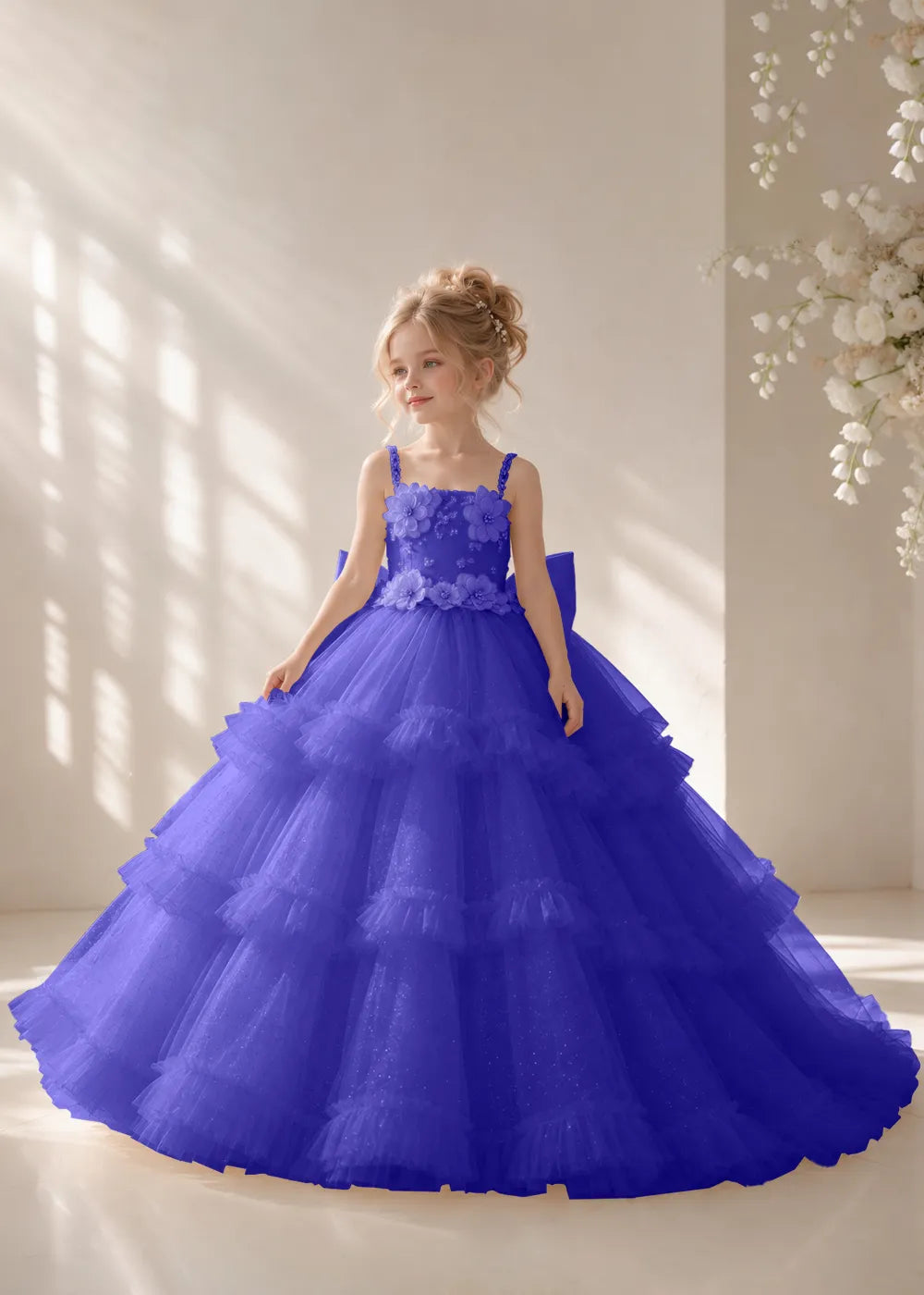 Front view of a child wearing a royal blue flower girl dress with beaded spaghetti straps, a bodice covered in three-dimensional fabric flowers and a multi-tiered tulle skirt.