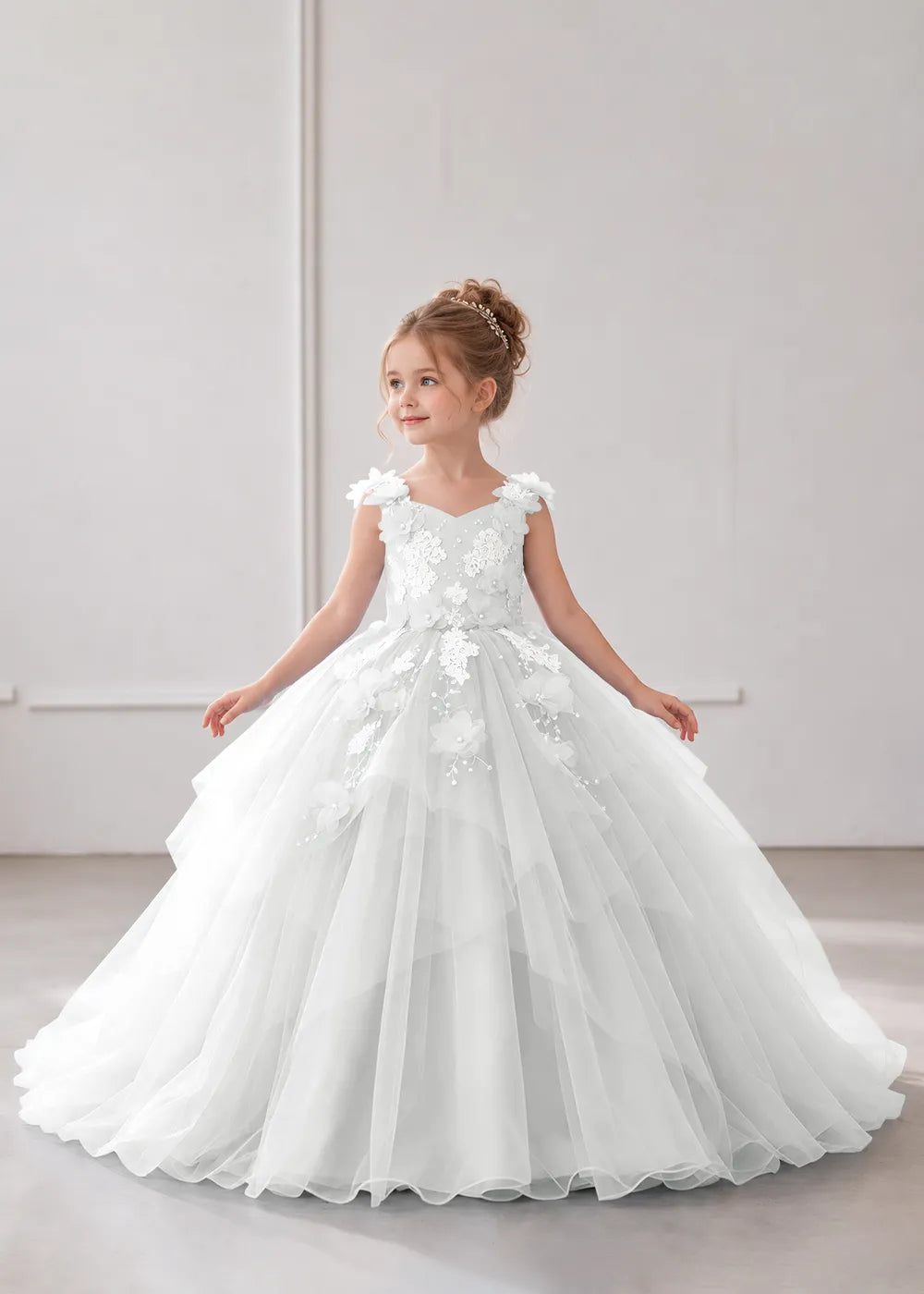 Front view of a child wearing a white flower girl dress with a V-neckline, floral shoulder appliques and lace and flower decoration across the bodice and hips.