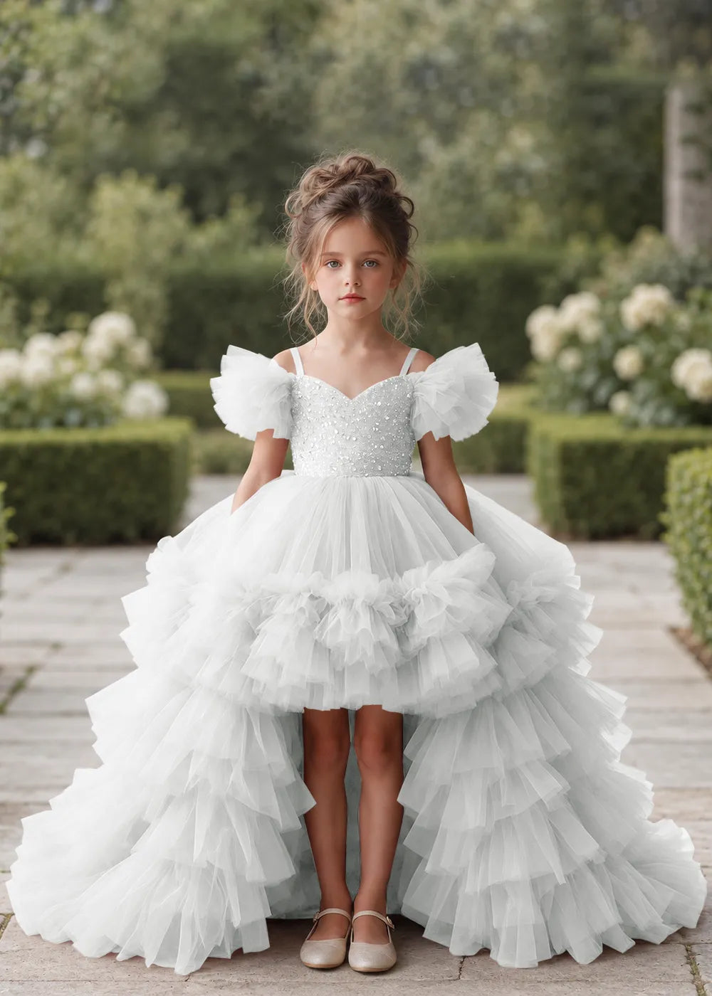 Front view of a child wearing a white flower girl dress with a V-neckline, a sequin-covered bodice and a tiered high-low tulle skirt with off-shoulder sleeve ruffles.