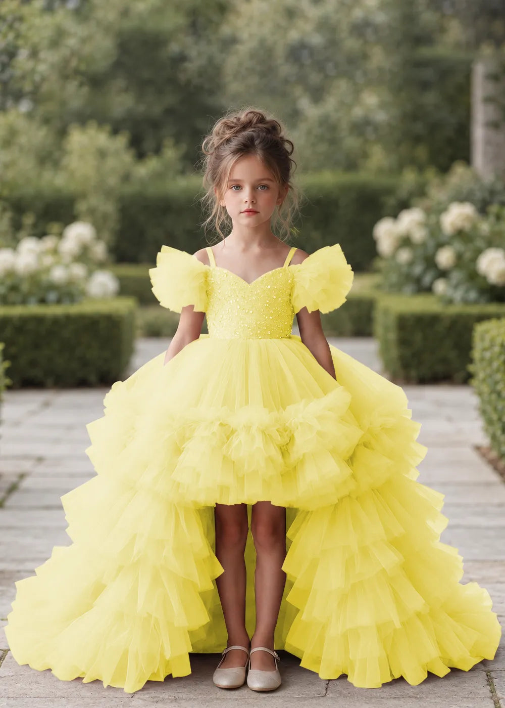 Front view of a child wearing a yellow flower girl dress with a V-neckline, a sequin-covered bodice and a tiered high-low tulle skirt with off-shoulder sleeve ruffles.